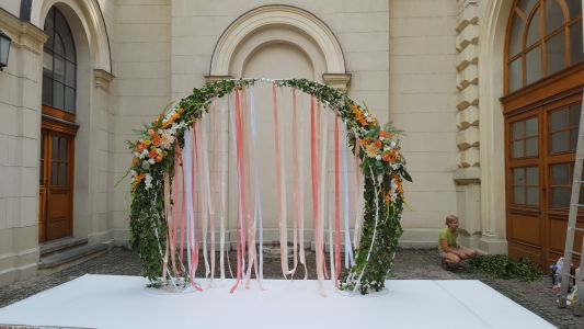 rundes Hochzeitstor mit Bändern und weiß-orangefarbenem Blumenarrangement (Rose, Dahlie, Gladiole)